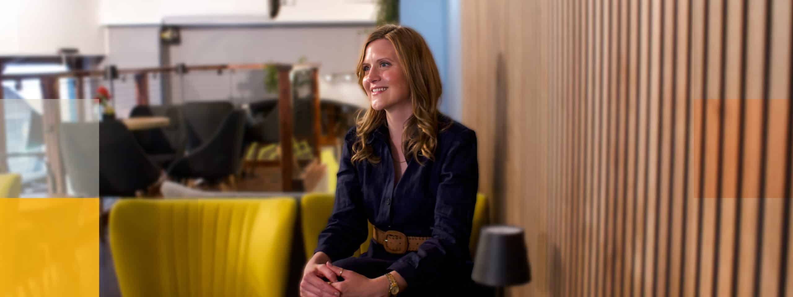 A person with long hair and a smile sits in a modern lounge area with yellow chairs and a wooden slatted wall, reminiscent of the contemporary spaces Desr&eacute; Hancocks might choose for Personal Coaching sessions. The background shows more seating and a railing, adding to the casual vibe.