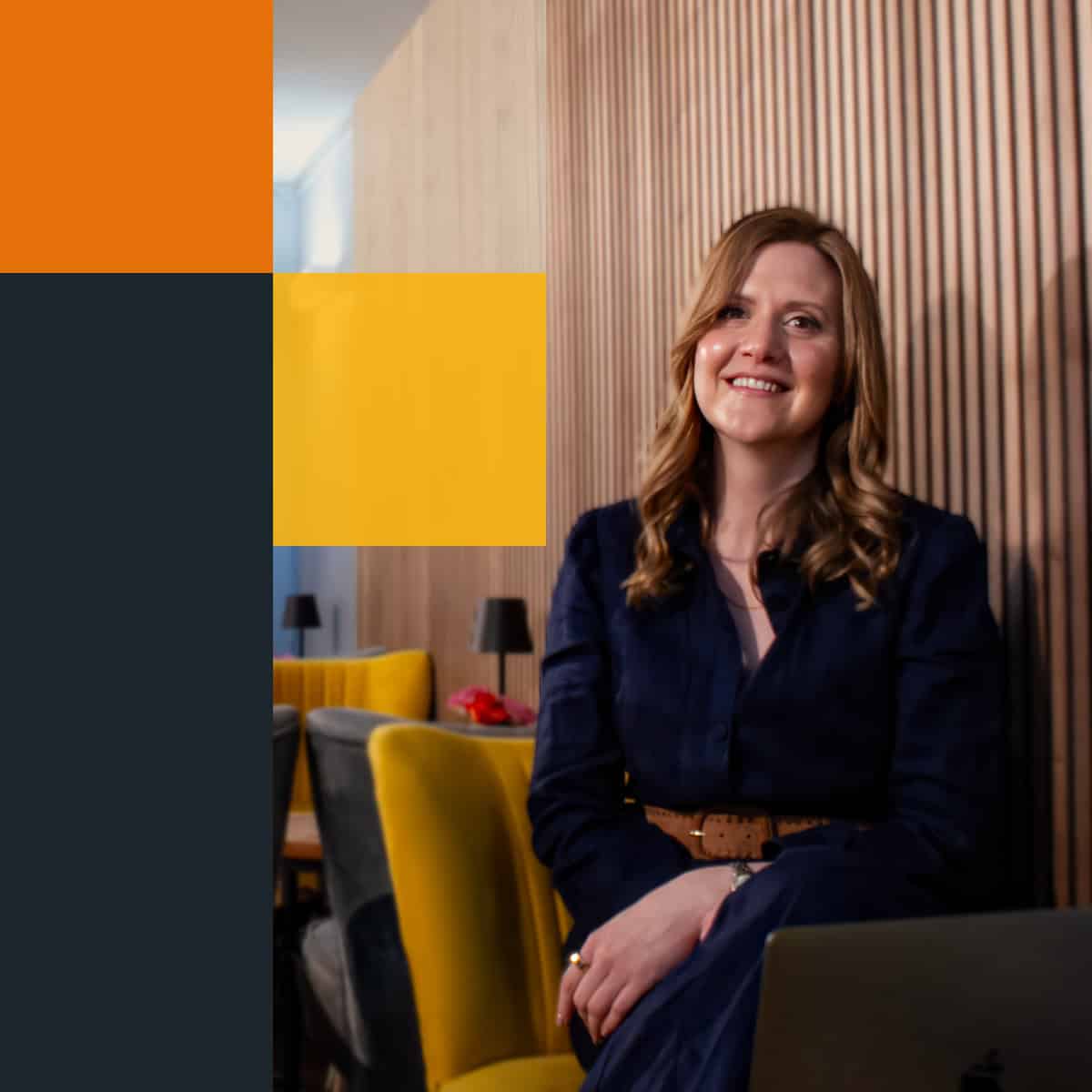 Desr&eacute; Hancocks seated in a modern office with a wooden accent wall and yellow chairs in the background. She is wearing a dark blue dress and has a laptop in front of her, representing Desr&eacute; Hancocks' Personal Coaching from Impact Stories Consulting.