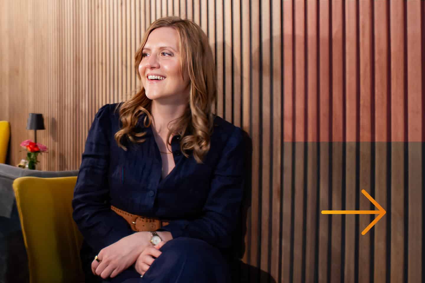 Desr&eacute; Hancocks sitting against a wooden panel wall. She is smiling and looking to her left. An orange arrow is drawn on the wall to the right of her, symbolizing the impactful guidance offered by Impact Stories Consulting.