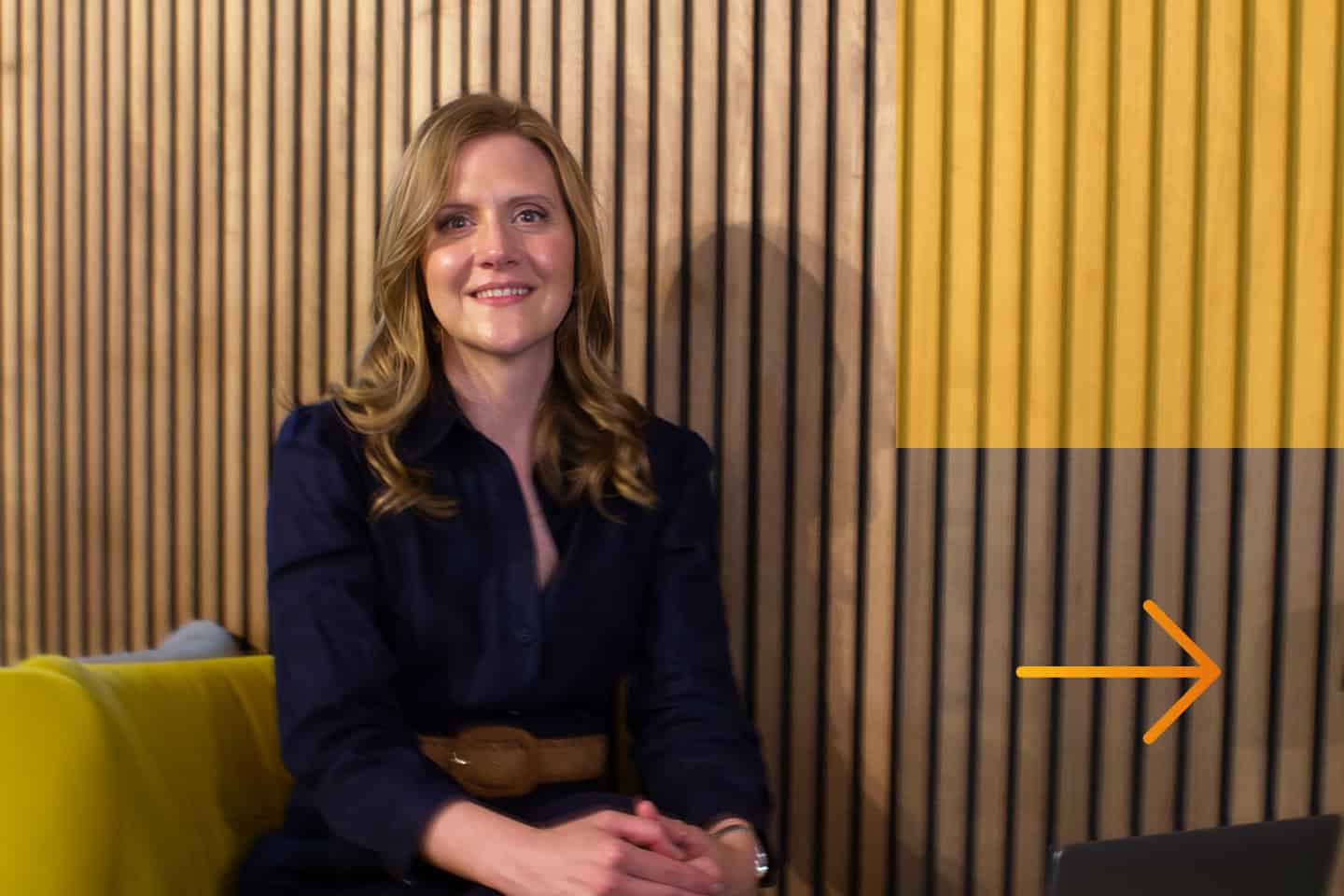 Desr&eacute; Hancocks sitting against a wooden panel wall. She is smiling and looking to her left. An orange arrow is drawn on the wall to the right of her, symbolizing the impactful guidance offered by Impact Stories Consulting.