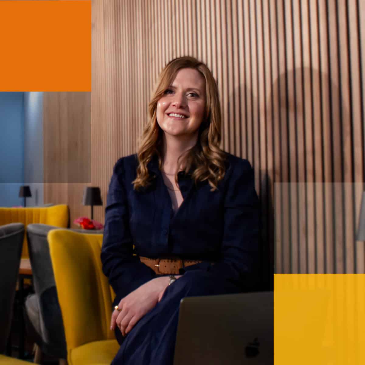 A woman with wavy blonde hair, wearing a navy blue dress, sits smiling with a laptop on her lap. She is in a modern room with wooden paneled walls, yellow chairs, and a dining table with lamps. The image has an overlay of orange geometric shapes.